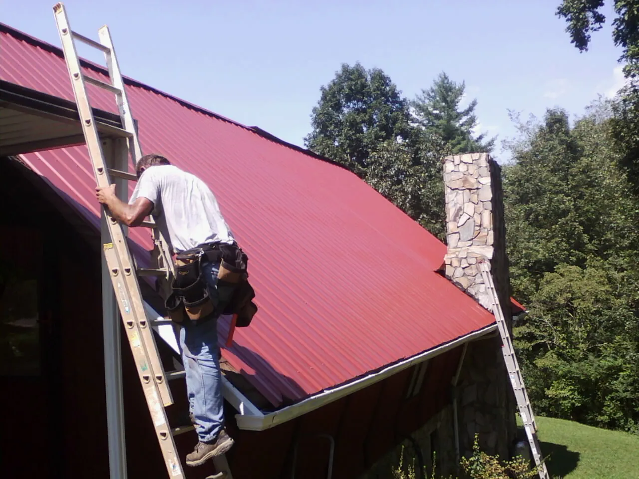 Roofer on a ladder beside a red metal roof and stone chimney — Soffit & Fascia Repair by Roofing Brothers in Macy